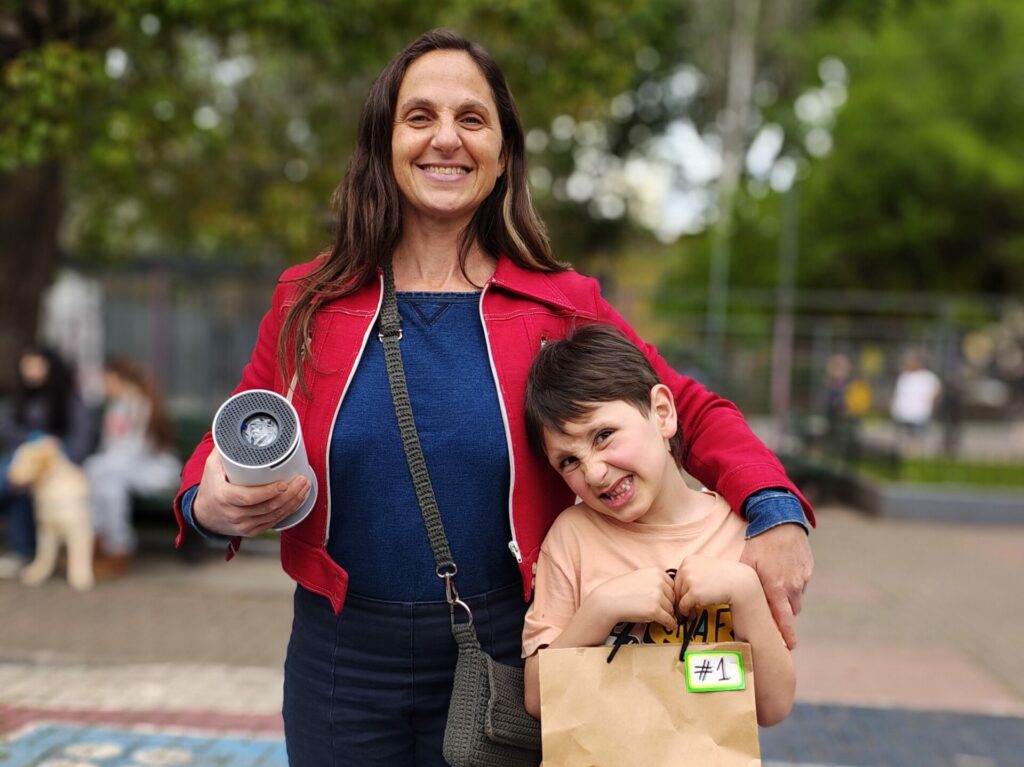 Madre e hijo en la plaza de la escuela, ella tiene el proyector en la mano, un tubo del tamaño de un rollo de cocina, él tiene la bolsa con el número 1 del premio.