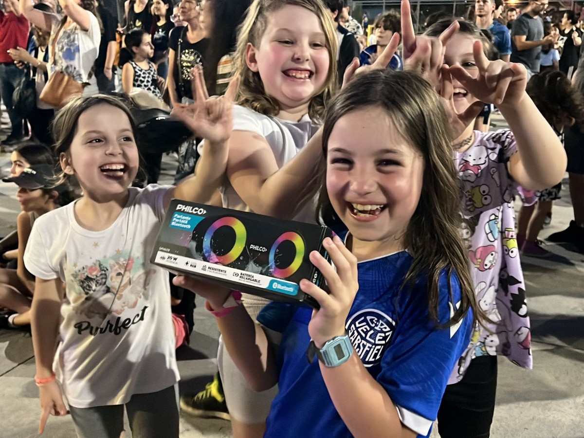 Una niña sostiene la caja de su parlante portátil, mientras sus amigas festejan alrededor, todas sonríen muy alegres.