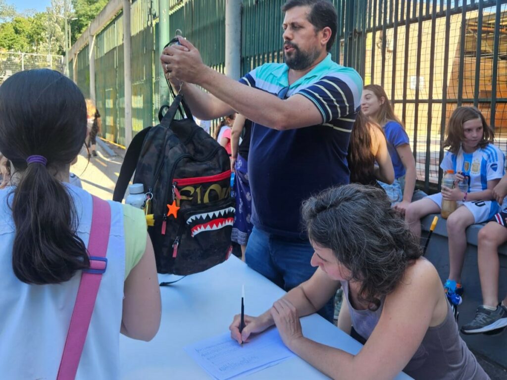 En la puerta de la escuela un papá pesa una mochila mientras una mamá toma nota, niños alrededor observan.