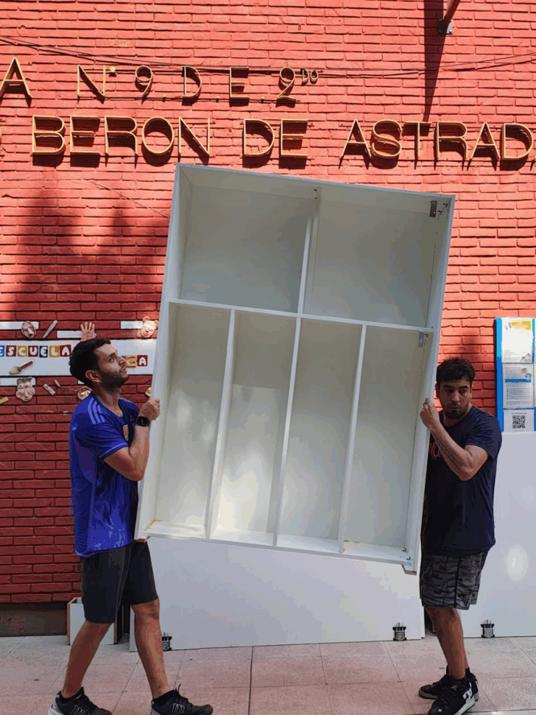 Dos hombres cargan un pesado mueble blanco frente a la entrada de la escuela.